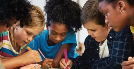 A group of young students standing over a desk in a classroom setting and writing on a poster.