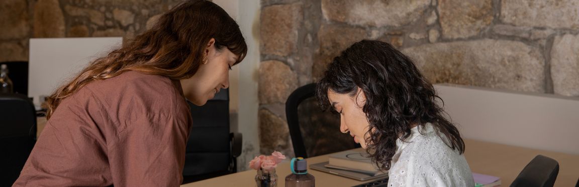 One person stands next to a second person sitting at a desk. Both individuals are looking down at the desk, focusing.